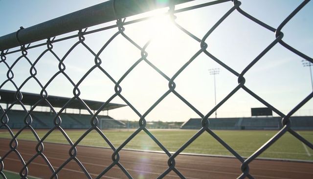 Backlit chain-link fence catching sunlight over track and empty stadium grandstand