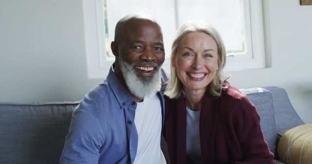 Mature interracial couple sitting on a couch in a well-lit living room, smiling at the camera. Ideal for advertising retirement plans, senior living, home lifestyle, health and wellness, and family-oriented products.