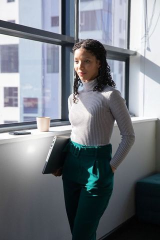 African american professional woman standing by sunlit office window holding laptop
