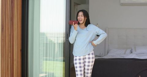 Laughing asian woman speaking into smartphone in bedroom doorway wearing pajamas