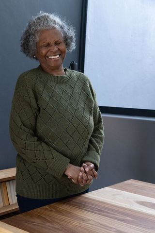 Joyful senior woman laughing by window in cozy green sweater