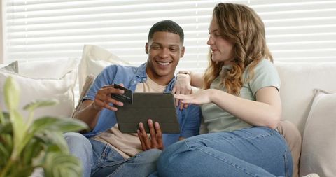 Diverse couple shopping online on tablet using credit card, relaxed cozy living room moment