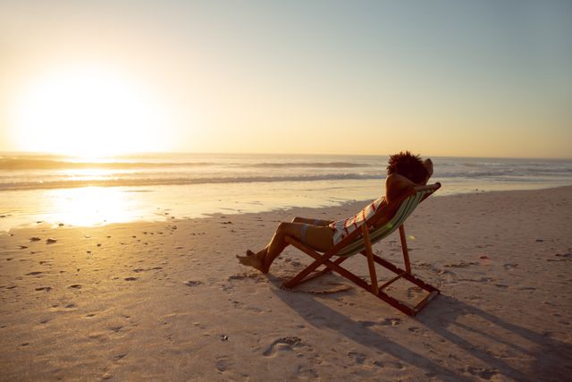 Man sitting in a beach chair with hands behind head, enjoying a serene sunset by the ocean. Ideal for themes of relaxation, vacation, leisure, and tranquility. Perfect for travel brochures, wellness blogs, and lifestyle magazines.