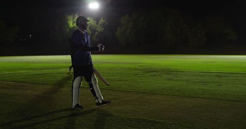 Night cricketer walking under floodlight carrying bat wearing helmet gloves and pads