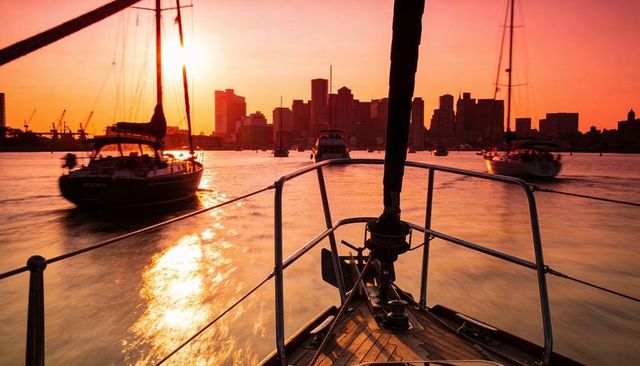Sailboat bow gliding through harbor at sunset with city skyline and golden reflections