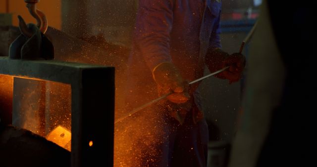 Worker is handling molten metal with protective gloves in an industrial facility. Bright orange glow and sparks indicate high temperature and intense heat. Ideal for content related to manufacturing processes, industrial safety, metallurgy, labor industry, and engineering. Useful for educational materials, industrial websites, or safety guideline publications.