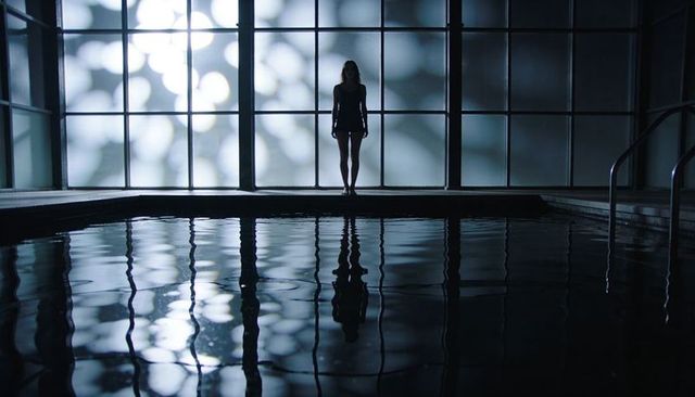 Silhouette woman standing at indoor pool edge reflecting bokeh lights and rippled water