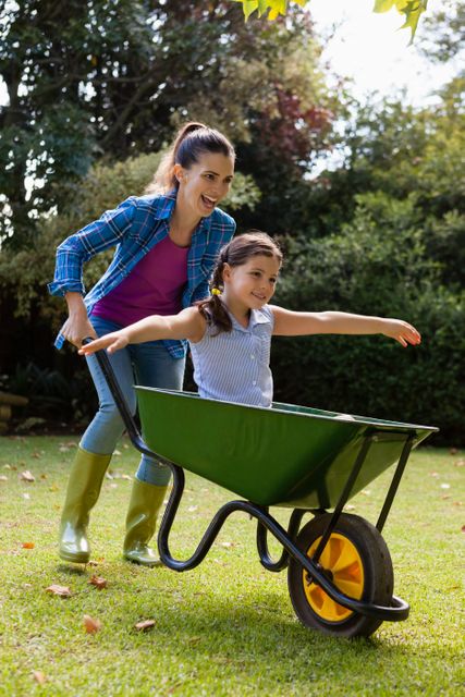 A cheerful mother is pushing her daughter who is sitting in a green wheelbarrow in a backyard. The scene is filled with joy and laughter, showcasing a playful moment between parent and child. This image is perfect for use in family-oriented advertisements, parenting blogs, outdoor activity promotions, and gardening websites.