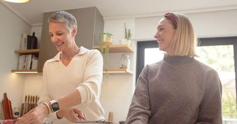 Mature mother and adult daughter cooking together in sunlit modern kitchen, smiling