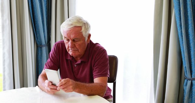 Elderly man sitting at table in casual attire, browses smartphone in bright living room near draped window. Useful for technology advertisements, elderly lifestyle magazines, and communication-themed projects.