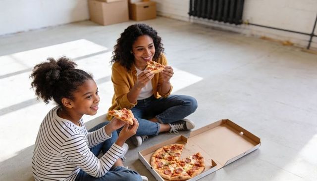 Mother and daughter eating pepperoni pizza on sunlit empty loft floor during move break