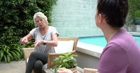 Senior woman and friend chatting poolside on wicker chairs, relaxed outdoor conversation