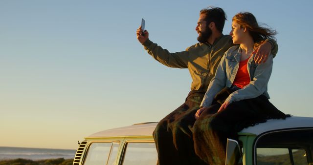 Young couple sitting on van roof, capturing selfie against beach during sunset. Ideal for depicting travel, adventure, road trips, and romantic getaways. Perfect for use in advertisements for travel agencies, outdoor lifestyle promotions, and social media content.