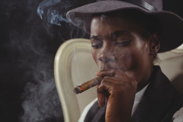 This image depicts an androgynous person wearing a hat and smoking a cigar while sitting in a vintage chair. The dark background and smoke create a moody and contemplative atmosphere. This image can be used in articles or advertisements related to lifestyle, relaxation, vintage fashion, or smoking culture.