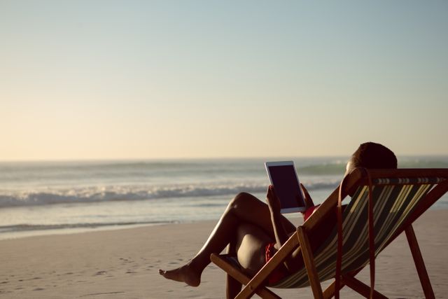 Woman sitting in a beach chair using a digital tablet by the ocean. Ideal for concepts related to summer vacations, remote work, digital nomad lifestyle, relaxation, and technology use in leisure time.