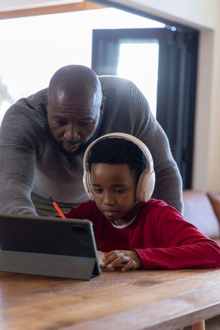 African american father guiding son on tablet wearing headphones for home learning