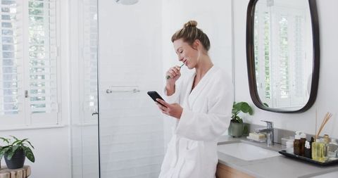 Woman in White Bathrobe Brushing Teeth and Using Smartphone