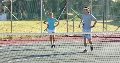 Mixed doubles partners practicing on sunny outdoor hard court with net rackets visors