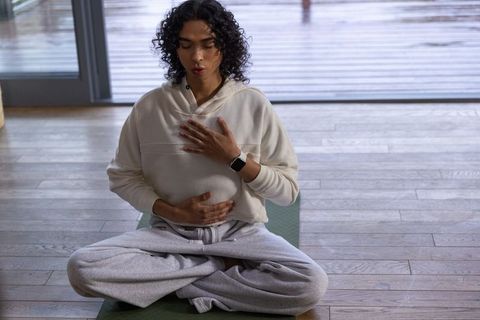Non-binary person meditating cross-legged on green mat by glass door in cream hoodie