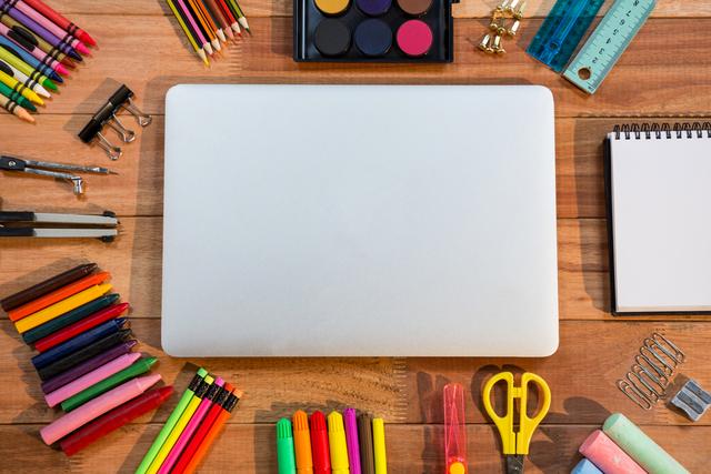 Laptop placed on wooden table surrounded by various colorful stationery items like pencils, markers, scissors, and notebooks. Ideal for use in educational materials, back-to-school promotions, office organization content, and creative workspace inspiration.