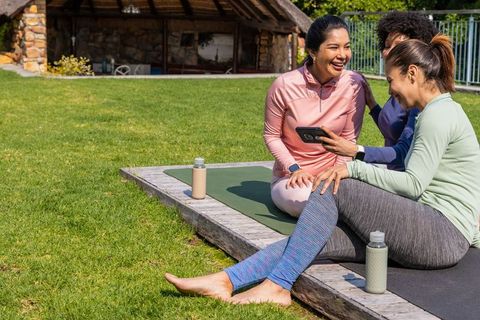 Diverse women laughing and sharing phone after yoga, sitting on deck mats in sunny park