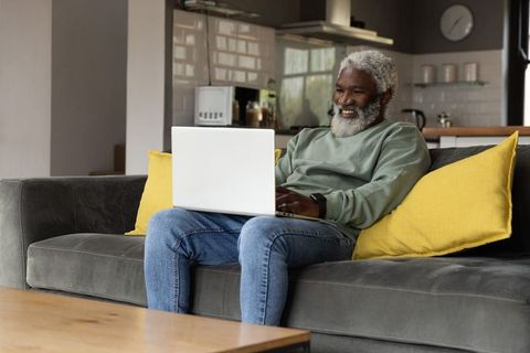 Senior african american man smiling and using laptop on sofa in cozy open-plan living room