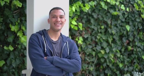 Smiling young man leaning on white column with ivy backdrop and casual hoodie