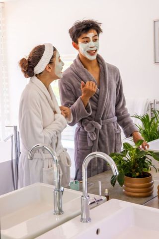 Diverse couple enjoying morning skincare routine with sheet masks and robes in bathroom