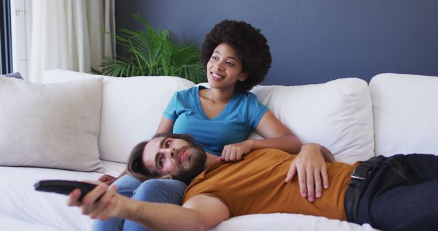 Multiracial couple relaxing on sofa watching TV, showcasing a comfortable home setting. They seem to be enjoying leisure time, holding remote control. Ideal for use in lifestyle articles, ads representing diverse relationships, or promotional material focusing on home entertainment.