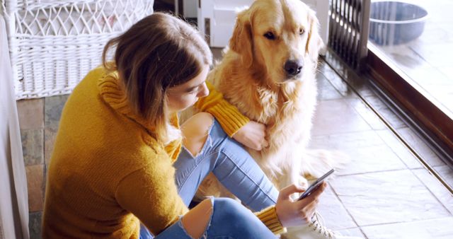 Young woman wearing a yellow sweater and jeans sitting on the floor next to a golden retriever dog, both looking at a smartphone. This image can be used for themes related to companionship, pet care, technology, everyday life, or online communication.