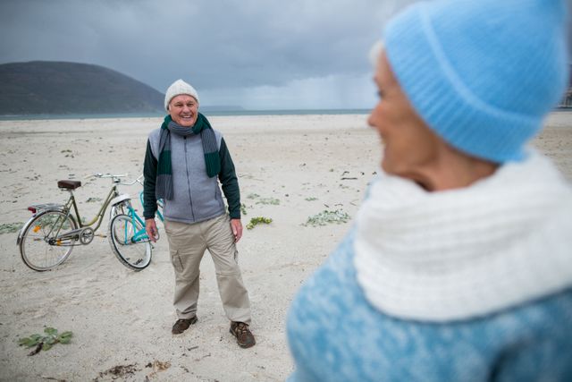 Senior couple enjoying a day at the beach with their bicycles. They are dressed in winter clothing, smiling and engaging in an active lifestyle. Ideal for use in advertisements promoting healthy living, retirement plans, travel, and outdoor activities for seniors.