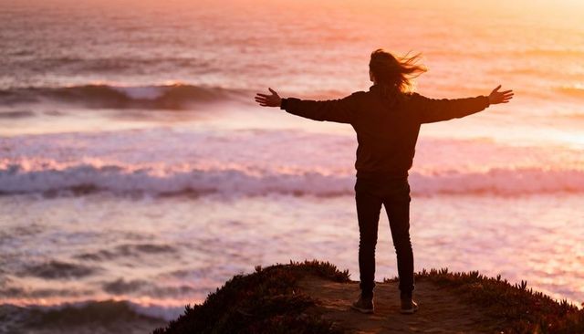 Standing silhouette spreading arms on coastal bluff at sunset overlooking ocean waves