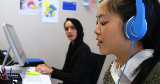 Image shows two office employees concentrating on work tasks. One person is wearing blue headphones and working on a computer, while the other is typing on another computer. This image is useful for illustrating collaborative work environments, modern office setups, and focus in the workplace. It can be used in articles about remote work, productivity tools, or teamwork in corporate settings.