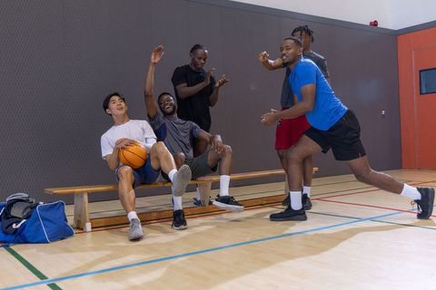 Courtside camaraderie: diverse men laughing and cheering while holding basketball on bench