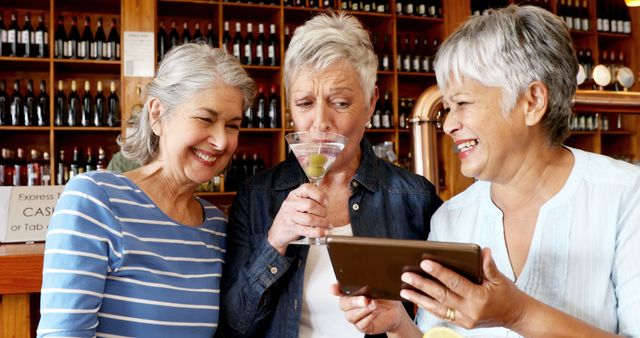 Senior women are at a bar, enjoying drinks and using a tablet. They are sharing laughter and happy moments together. This photo is ideal for use in advertisements related to retirement communities, technology for seniors, social gatherings, or lifestyle content aimed at an older demographic.
