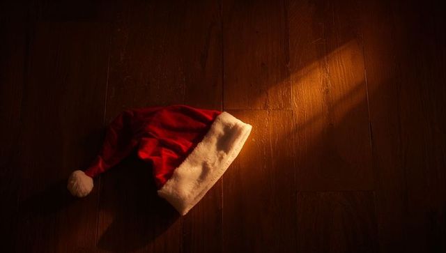 Red santa hat catching warm beam on hardwood floor, crumpled velvet fur pom-pom and shadow