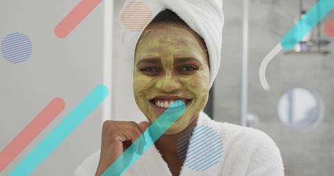 Smiling woman wearing green facial mask and towel turban brushing teeth in bathroom