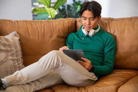 Asian man relaxing on tan leather couch using tablet while wearing headphones and smartwatch