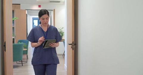 Female nurse walking clinic corridor using tablet and stylus in navy scrubs with id badge