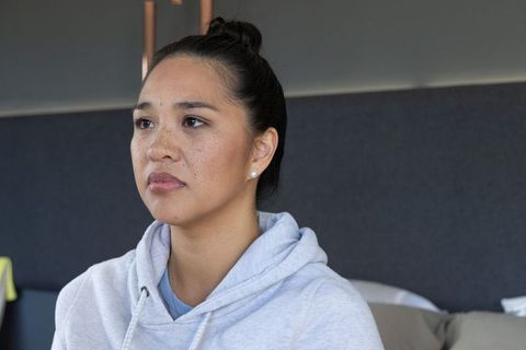 Thoughtful asian woman wearing light gray hoodie sitting in minimalist bedroom calm portrait