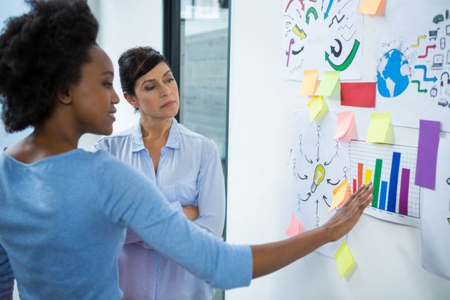 Two female designers are collaborating in a creative office, discussing project ideas and pointing at sticky notes and charts on a glass wall. This image can be used to illustrate teamwork, brainstorming sessions, and the creative process in a professional setting.