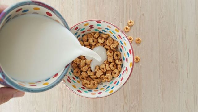 Polka-dot bowl and pitcher pouring milk over oat ring cereal on light wood tabletop