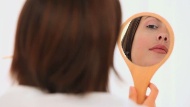 A woman with brown hair is examining her reflection in a handheld mirror. This scene evokes themes of self-reflection and beauty. Ideal for beauty care, self-awareness concepts, and lifestyle content related to personal grooming.