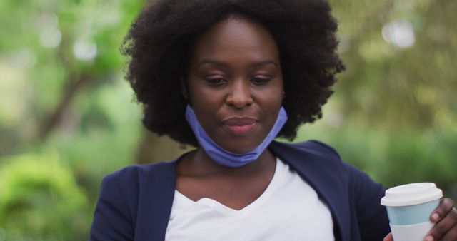 African American woman with afro hair holding takeaway coffee, wearing a face mask. The background is green and blurred, suggesting a park or garden setting. Ideal for illustrating themes of health, pandemic practices, outdoor activities, and everyday life.