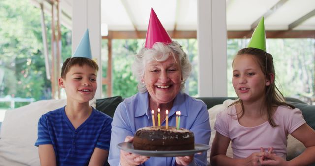 Caucasian senior woman in party hat blowing candles on birthday cake while her grandchildren sitting besides her on couch at home. social distancing during coronavirus quarantine lockdown.