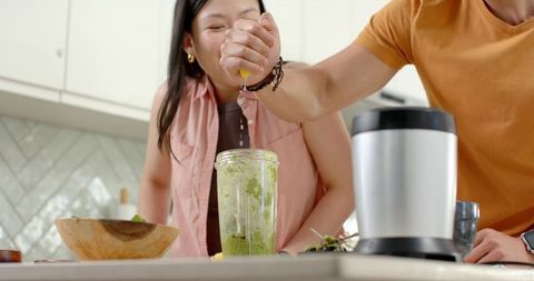 Asian couple squeezing lemon and making green smoothie together in bright modern kitchen