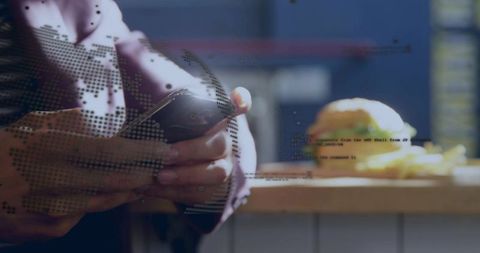 Tapping smartphone at cafe counter with burger and fries, digital code and world map overlay