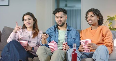 Friends watching movie on couch, sharing popcorn and bottled drinks for movie night