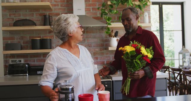 Happy senior african american husband giving flowers to biracial wife in kitchen. Romance, love, senior lifestyle, togetherness and domestic life.