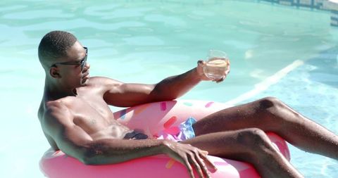 African american man reclining on pink donut float in sunny pool sipping drink enjoying leisure